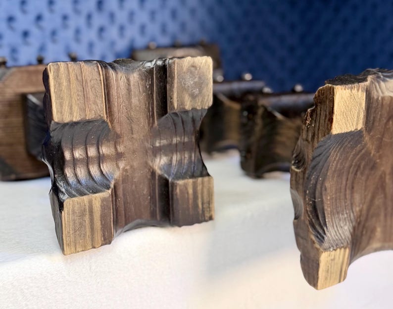 May include: A collection of dark brown wooden blocks with a square, carved cross-shaped center and textured, grooved edges. The wood grain is visible, showing a mix of deep espresso and lighter tan tones across the geometric, blocky surfaces.