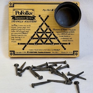 May include: A rectangular wooden PoFolks Signature Series Triangle Solitaire game board with a black circular peg holder and printed instructions. The board features a triangular grid of holes and includes a collection of dark metal nail-style pegs.