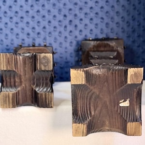 May include: Two dark brown wooden candle holders with a square base and a textured, carved surface. The square bases feature a cross-shaped indentation in the center, and one base has a small, rectangular white sticker attached to the wood surface.