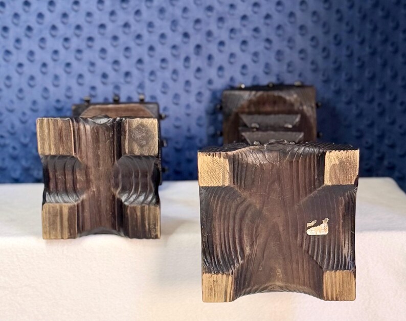 May include: Two dark brown wooden candle holders with a square base and a textured, carved surface. The square bases feature a cross-shaped indentation in the center, and one base has a small, rectangular white sticker attached to the wood surface.