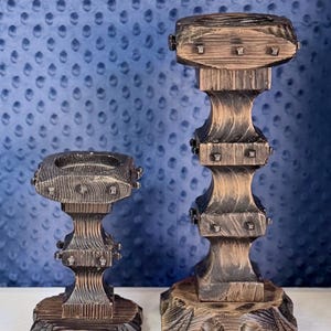 May include: Two dark brown wooden candle holders with a distressed finish and visible wood grain. Each holder features a square top, a tiered central column, and a wide base, all accented with small, evenly spaced metal studs on the edges.