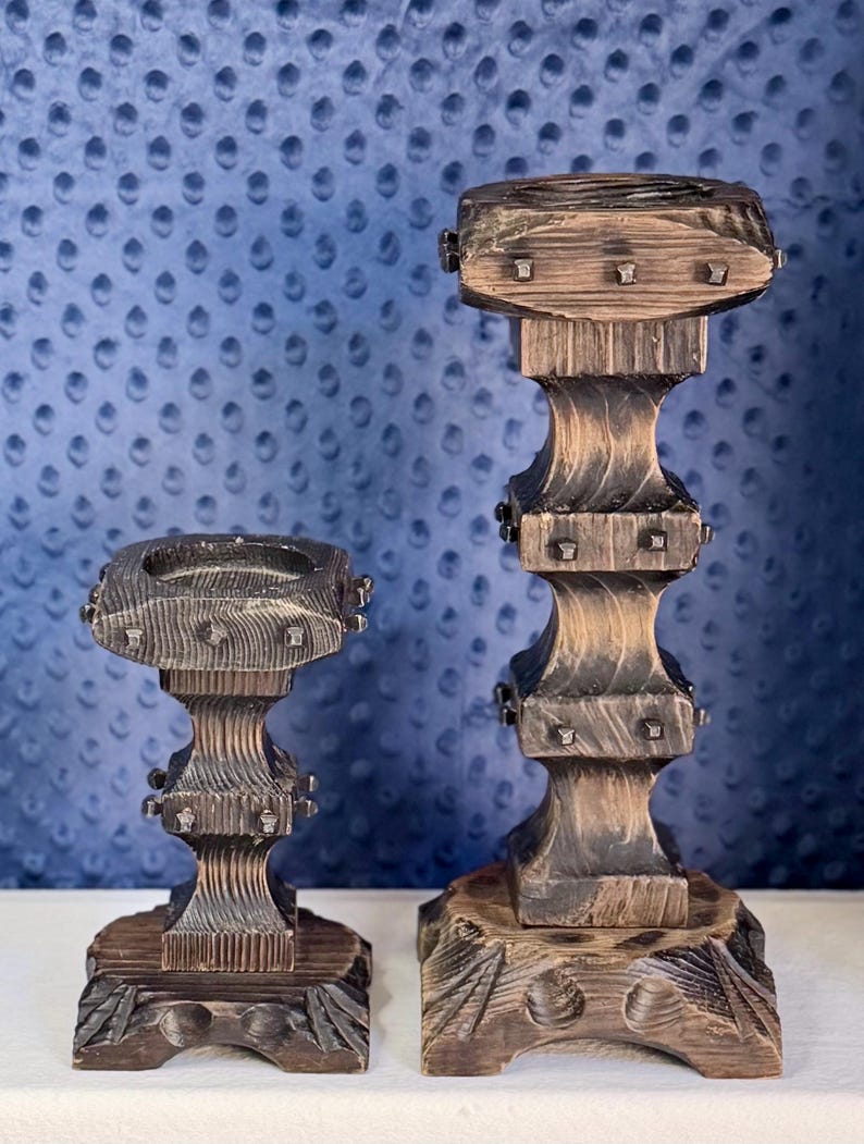 May include: Two dark brown wooden candle holders with a distressed finish and visible wood grain. Each holder features a square top, a tiered central column, and a wide base, all accented with small, evenly spaced metal studs on the edges.