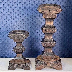 May include: Two dark brown wooden candle holders with a textured, weathered finish. The shorter holder has a square base and a single central section, while the taller holder features a square base and two stacked sections, both accented with small metal studs.