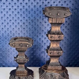 May include: Two dark brown wooden candle holders with a textured, weathered finish. Each holder features a square top, a multi-tiered central column, and a wide, carved base. Small, square metal studs are embedded into the sides of the top sections.