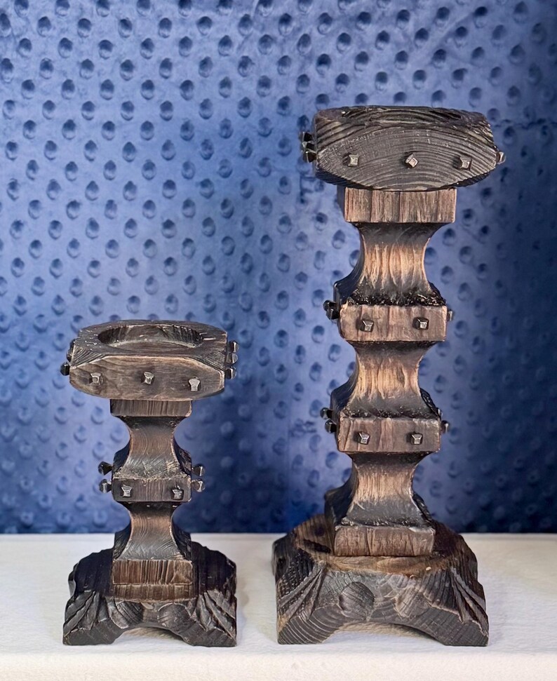 May include: Two dark brown wooden candle holders with a textured, weathered finish. Each holder features a square top, a multi-tiered central column, and a wide, carved base. Small, square metal studs are embedded into the sides of the top sections.