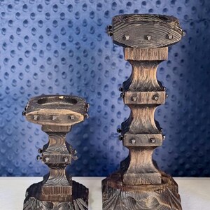 May include: Two dark brown wooden candle holders with a scorched finish and square bases. Each holder features a tiered, carved design with small metal studs embedded along the edges of the square platforms and a circular indentation on top.