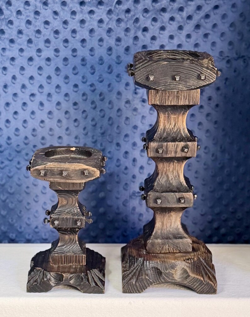May include: Two dark brown wooden candle holders with a scorched finish and square bases. Each holder features a tiered, carved design with small metal studs embedded along the edges of the square platforms and a circular indentation on top.