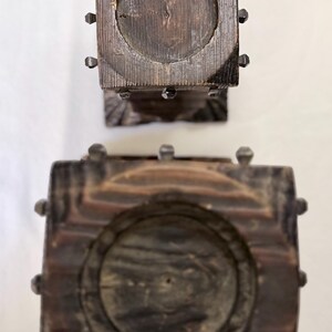 May include: Two square dark brown wooden candle holders viewed from directly above. Each holder features a circular recessed center for a candle and is decorated with small, dark metal studs along the edges of the square wooden frame.