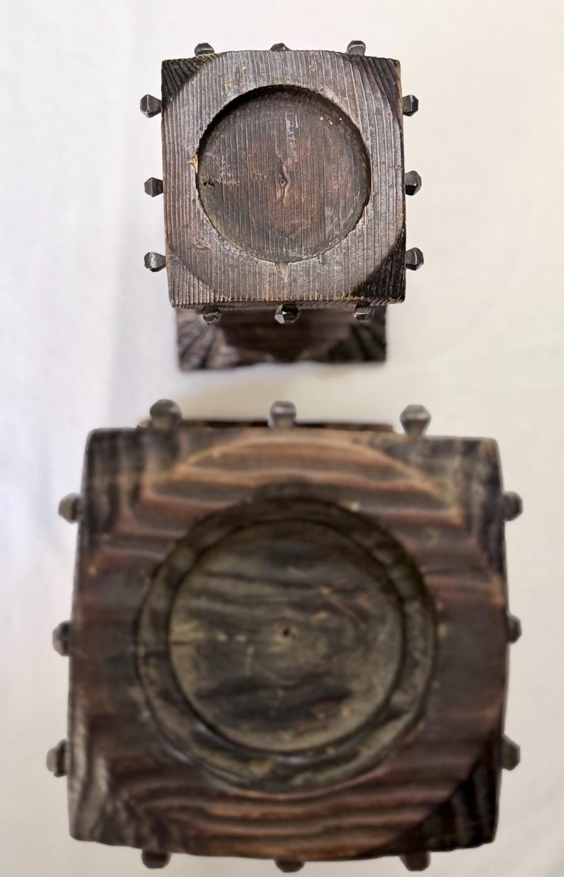 May include: Two square dark brown wooden candle holders viewed from directly above. Each holder features a circular recessed center for a candle and is decorated with small, dark metal studs along the edges of the square wooden frame.