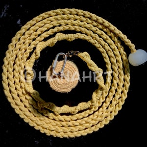 Könnte beinhalten: Ein gelbes geflochtenes Seil mit einem Karabinerhaken aus Metall und einem weißen runden Objekt daran. Das Seil ist zu einem Kreis gewickelt.