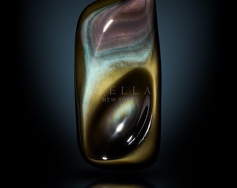 May include: A polished, oblong gemstone with swirling patterns of brown, gold, blue, and purple. The stone has a glossy finish and is set against a dark background. The word "STELLA NEW YORK" is printed on the stone.