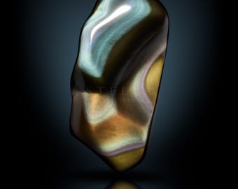 May include: A polished, iridescent stone with a smooth, organic shape. The stone features a blend of colors, including brown, gold, blue, and purple, with a glossy finish. The word "STELLA" is at the bottom.