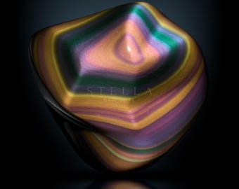 May include: A polished, iridescent sculpture with a smooth, rounded form. The object features bands of color including gold, green, purple, and orange. The word "STELLA NEW YORK" is printed on the object. The word "STELLA" is printed at the bottom.