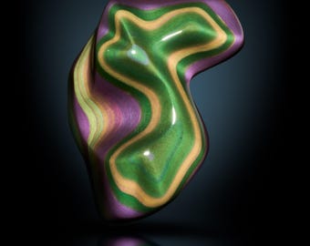 May include: A glossy, abstract sculpture with a flowing, organic shape. The piece features layers of green, purple, and gold tones. The word "STELLA" is printed at the bottom of the image.