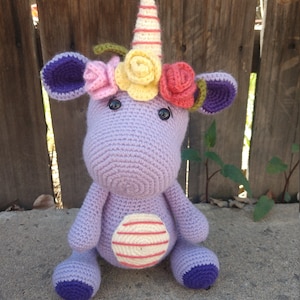 May include: A crocheted purple unicorn toy with a pink, yellow, and red flower crown. The unicorn has a white and pink striped belly and purple hooves.