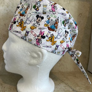 May include: White surgical scrub cap featuring a repeating pattern of Disney characters dressed as medical professionals. Mickey Mouse, Minnie Mouse, Donald Duck, and Pluto are depicted alongside medical tools and prescription bottles. The cap has a tie in the back.