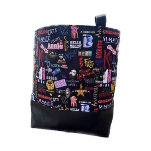 May include: Black tote bag with a colorful print of Broadway show titles, including "Chicago", "Cats", "Summer", "Grease", "Annie", "Hello Dolly", "Rent", "Wicked", "Hamilton", "Phantom of the Opera", "Anastasia", and "A Chorus Line".