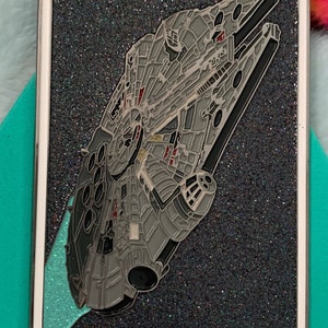 May include: A silver and black enamel pin featuring the Millennium Falcon from Star Wars. The pin is shaped like a tarot card with the Roman numeral VII and the text "The Chariot" on it.