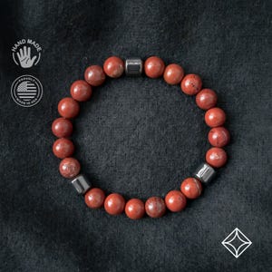 Op de afbeelding: Rode jaspis kralenarmband met drie zilverkleurige metalen kubusaccenten. De armband heeft ronde, gepolijste rode jaspis kralen. De achtergrond is een donker, getextureerd oppervlak. De afbeelding bevat ook een "Hand Made" en "Proudly Made in USA" logo.