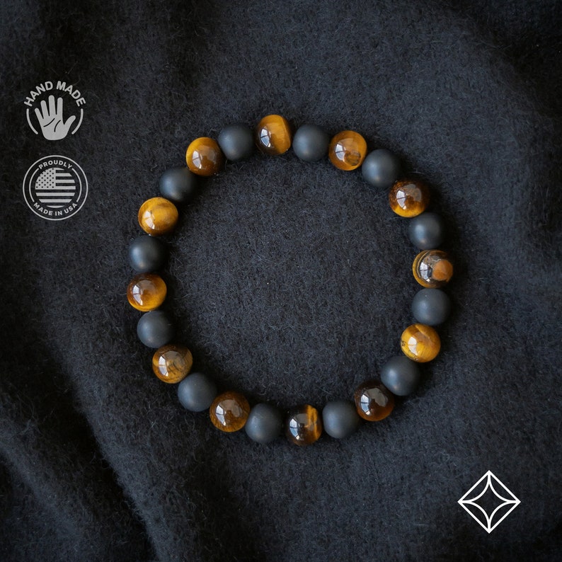Peut inclure: Bracelet fait &agrave; la main avec des perles noires mates et des perles &oelig;il de tigre altern&eacute;es. Les perles &oelig;il de tigre sont d'un riche brun dor&eacute;. Le bracelet est pr&eacute;sent&eacute; sur une surface sombre et textur&eacute;e. Comprend un logo "Hand Made" et "Proudly Made in USA".