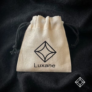 Peut inclure: Un petit sac beige &agrave; cordon de serrage avec un logo en forme de losange noir et le mot "Luxane" imprim&eacute; en dessous. Le sac a une fermeture &agrave; cordon noire et est pos&eacute; sur un fond sombre et textur&eacute;.