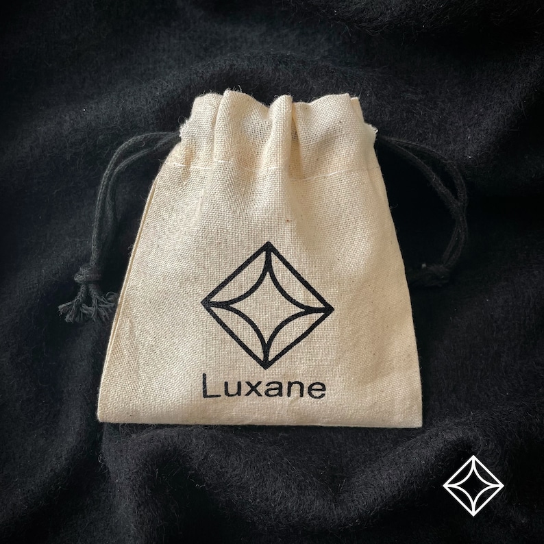 Peut inclure: Un petit sac beige &agrave; cordon de serrage avec un logo en forme de losange noir et le mot "Luxane" imprim&eacute; en dessous. Le sac a une fermeture &agrave; cordon noire et est pos&eacute; sur un fond sombre et textur&eacute;.