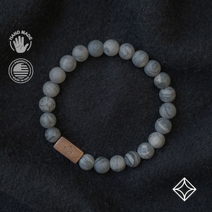 Puede incluir: Un brazalete de cuentas de piedra gris con un dije rectangular de cobre con un diseño geométrico. El brazalete está sobre un fondo de tela negro.