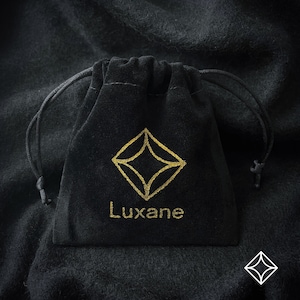 May include: A black velvet pouch with a gold drawstring closure. The pouch has a gold logo with a diamond shape and the word "Luxane" printed below it.