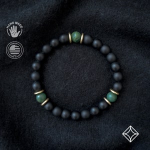 May include: A black matte beaded bracelet with two green gemstone beads and gold accents. The bracelet is made in the USA.