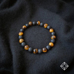 Peut inclure: Un bracelet de perles alternant des pierres noires mates et des pierres &oelig;il de tigre dor&eacute;es. Le bracelet est pr&eacute;sent&eacute; sur un fond sombre et textur&eacute;, soulignant le contraste entre les couleurs et les textures des perles.
