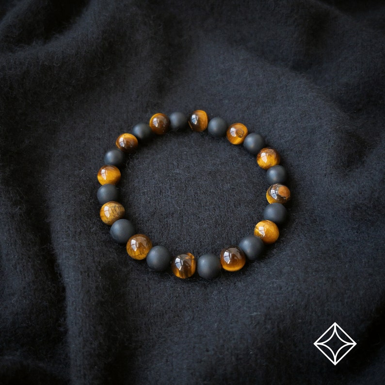 Peut inclure: Un bracelet de perles alternant des pierres noires mates et des pierres &oelig;il de tigre dor&eacute;es. Le bracelet est pr&eacute;sent&eacute; sur un fond sombre et textur&eacute;, soulignant le contraste entre les couleurs et les textures des perles.