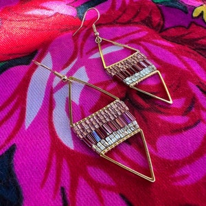 May include: A pair of gold triangle earrings with pink and silver beads. The earrings are on a pink and purple floral fabric background.