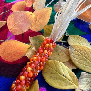 May include: A beaded corn cob with orange and yellow beads, lying on a colorful fabric with fall leaves.