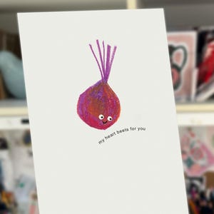 May include: A white greeting card with a cartoon beet illustration. The beet is pink and purple with a smiling face and purple stems. The text "my heart beets for you" is printed below the beet. The card is held in a hand.