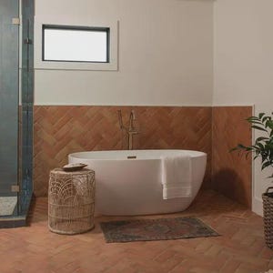May include: A modern bathroom with a white oval bathtub, a woven side table, and a small patterned rug. The walls are a warm terracotta color, and the floor is tiled in a matching brick pattern. A white towel hangs on the tub.