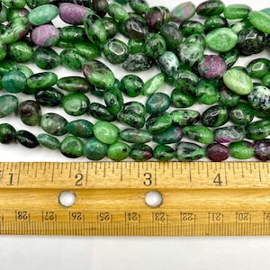 May include: A strand of green and pink gemstone beads, approximately 1 inch in length, laid out on a wooden ruler.