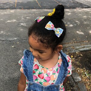 May include: A young girl with dark brown hair wearing a blue denim overall dress with a white shirt underneath. The shirt has a floral pattern with strawberries and peaches. The girl has three floral patterned hair bows in her hair.
