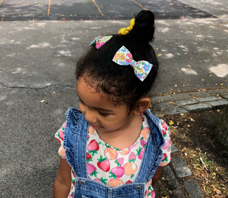 May include: A young girl with dark brown hair wearing a blue denim overall dress with a white shirt underneath. The shirt has a floral pattern with strawberries and peaches. The girl has three floral patterned hair bows in her hair.