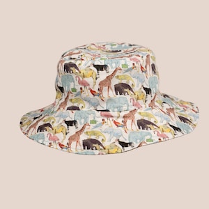 May include: A white bucket hat with a colorful animal print featuring giraffes, elephants, flamingos, and other wildlife. The hat has a wide brim and a rounded crown, suitable for sun protection.