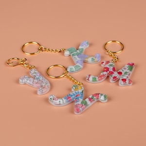 May include: Five floral letter keyrings. The letters are in the shape of the letters A, K, N, and two Ys. Each letter has a gold-coloured chain and keyring. The letters are white with a colourful floral pattern.