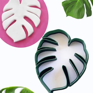May include: A white monstera leaf-shaped clay piece on a pink background, next to a green plastic leaf-shaped cookie cutter. A realistic green monstera leaf is also visible. The items are set against a white background.