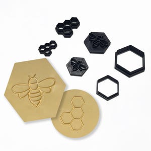 May include: A collection of bee-themed craft tools. Includes a large tan hexagon and circle with bee and honeycomb designs, black hexagon and honeycomb shapes, and black bee stamps. Ideal for crafting and DIY projects.