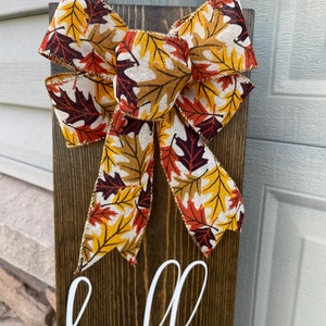 May include: A wooden sign with the word "hello" written in white paint. The sign is decorated with a large bow made of fabric with a fall leaf pattern in shades of orange, yellow, and brown.