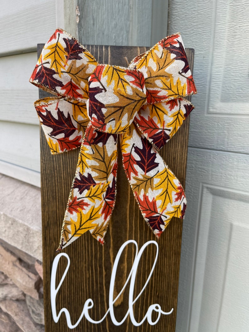 May include: A wooden sign with the word "hello" written in white paint. The sign is decorated with a large bow made of fabric with a fall leaf pattern in shades of orange, yellow, and brown.