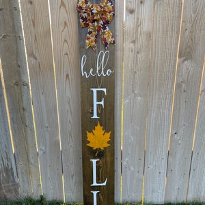 May include: Wooden sign with the words "hello fall" in white lettering. The sign is decorated with a brown and yellow bow and a yellow leaf.