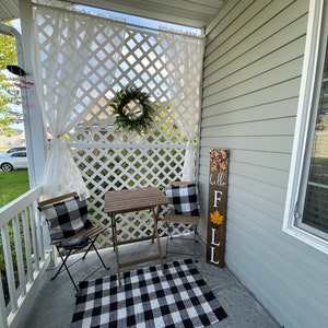 May include: A black and white checkered rug sits on a concrete porch with a small wooden table and two black metal chairs. A white lattice wall with white sheer curtains is behind the table. A wooden sign with the text "hello fall" is leaning against the wall.