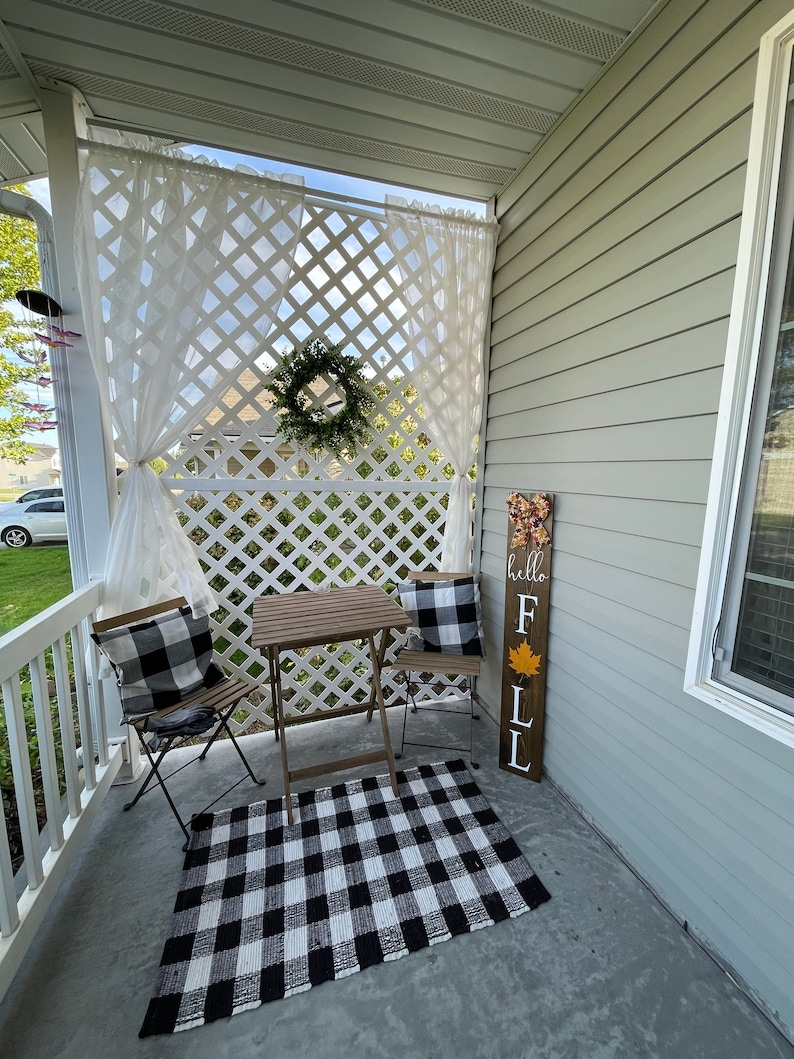 May include: A black and white checkered rug sits on a concrete porch with a small wooden table and two black metal chairs. A white lattice wall with white sheer curtains is behind the table. A wooden sign with the text "hello fall" is leaning against the wall.