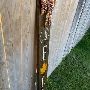 May include: Wooden sign with the words "hello fall" and a fall leaf design. The sign is decorated with a brown and white bow with a fall leaf pattern.