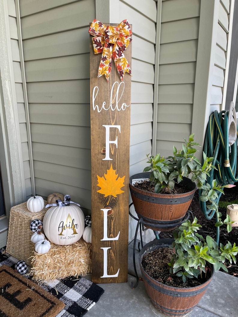 May include: A wooden sign with the words "hello fall" and a large orange leaf. The sign is decorated with a fall-themed bow and is displayed on a porch with pumpkins and other fall decorations.