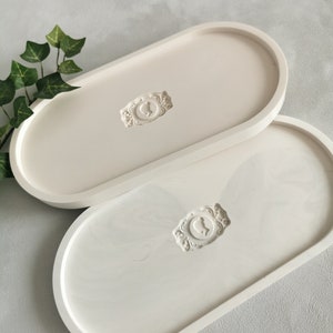 Peut inclure: Deux plateaux ovales blancs avec un motif en relief au centre. Le motif est un cercle avec un oiseau stylisé à l'intérieur. Les plateaux sont faits d'un matériau lisse et mat.